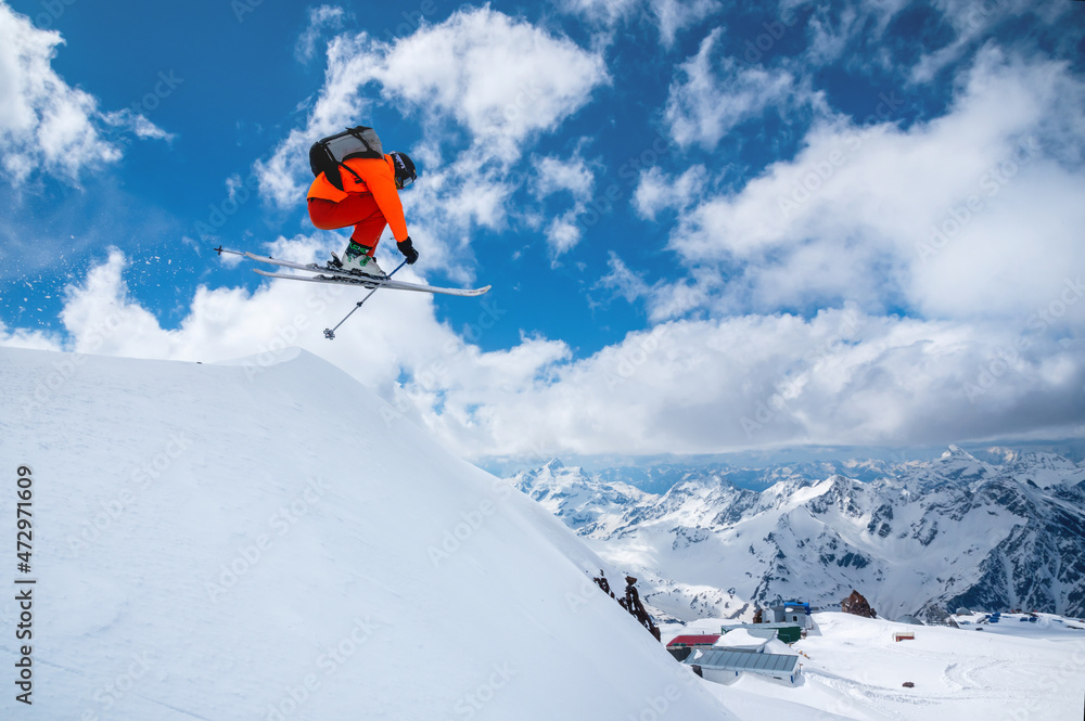 A professional skier in an orange suit jumps from a high cliff against a background of blue sky and clouds, leaving a trail of snow powder in the mountains.
