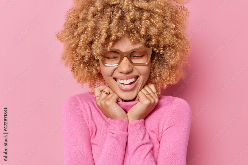 Happy positive young woman smiles broadly feels amused keeps hands on collar of jumper recalls something funny wears eyeglasses stands amused isolated over pink background. Emotions concept. Happy positive young woman smiles broadly feels amused keeps hands on collar of jumper recalls something funny wears eyeglasses stands amused isolated over pink background. Emotions concept.