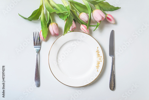 Spring holiday place setting; Top view of white plate, fork and knife and bouquet of pink tulips above