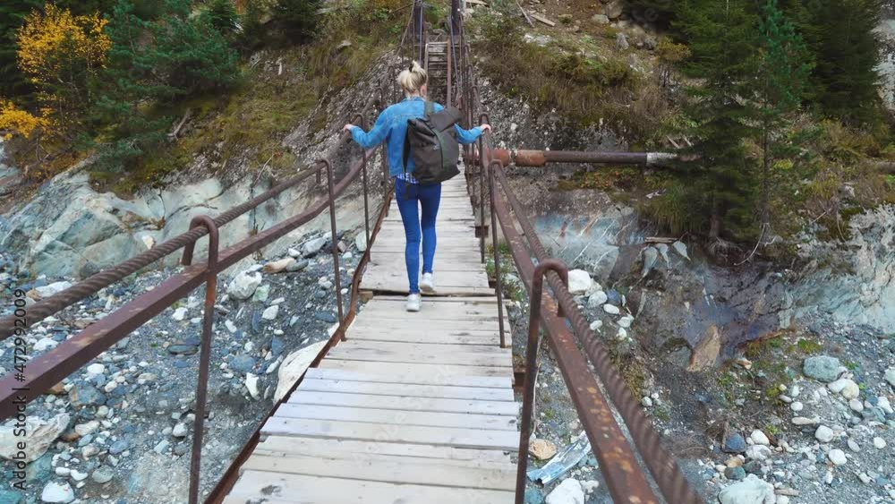 young beautiful slender woman in a denim suit walking on a suspension bridge alone in picturesque place across a stormy mountain river. She's scared, upstairs. Mountains in autumn. Chalaadi glacier. young beautiful slender woman in a denim suit walking on a suspension bridge alone in picturesque place across a stormy mountain river. She's scared, upstairs. Mountains in autumn. Chalaadi glacier.