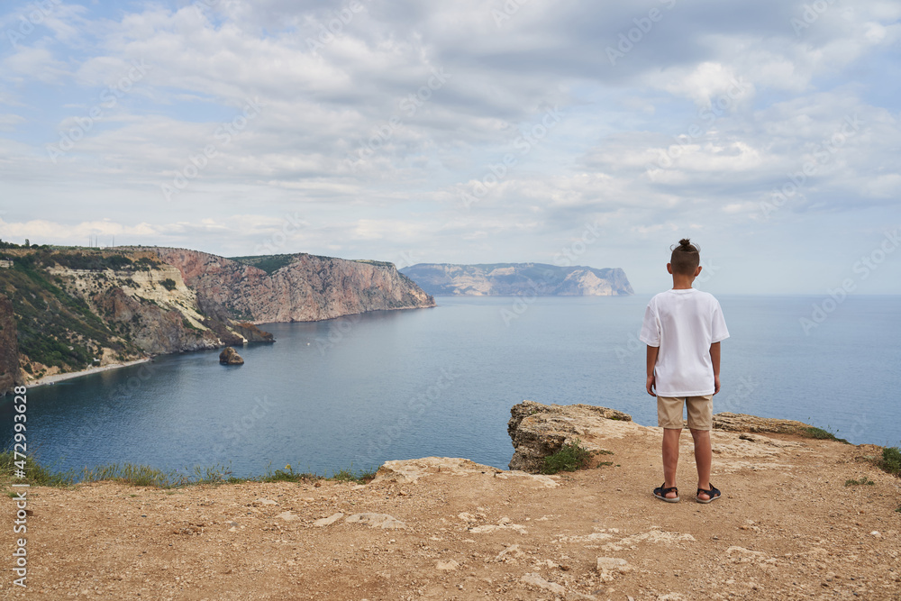 a boy in light clothes against the background of a beautiful seascape stands and looks at the rocky coastline, stretching into the distance, view from the back.