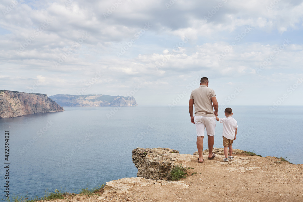 young man and little son stand by the hand on the edge of the rocky coast with a beautiful sea view. back view