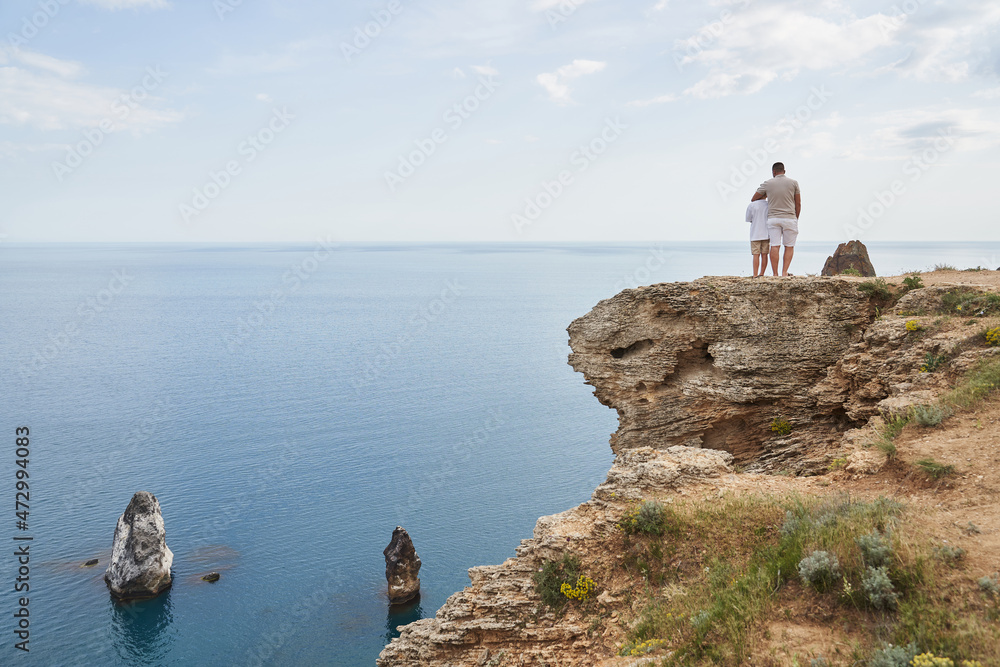 dad and son are standing on a rocky sea shore. dad hugging his son, view from the back
