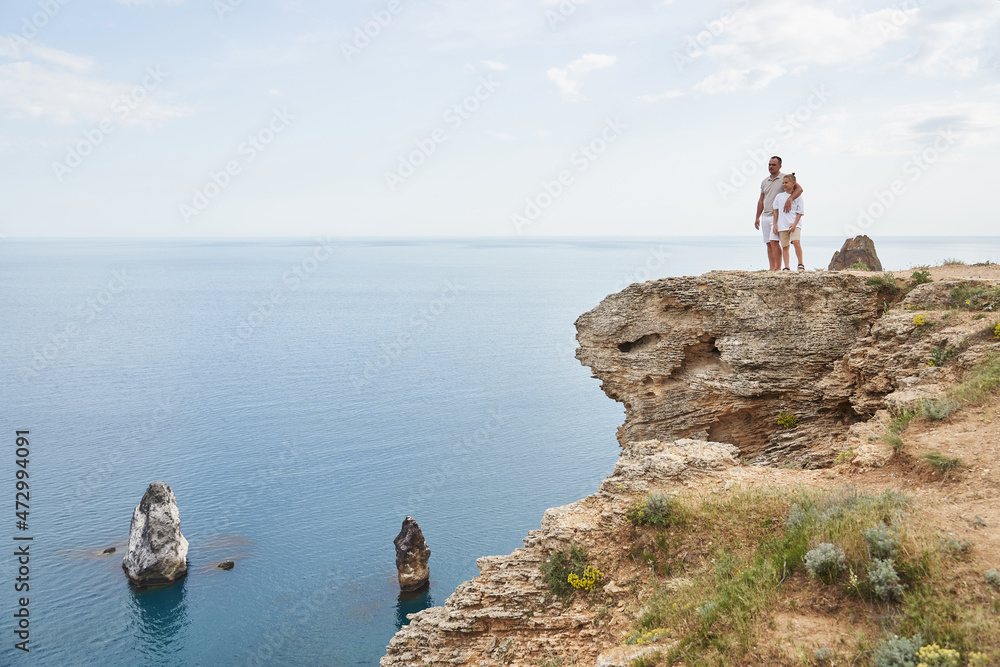 dad and boy are standing on the rocky coast of the sea. dad hugs his son by the shoulders and they look into the distance