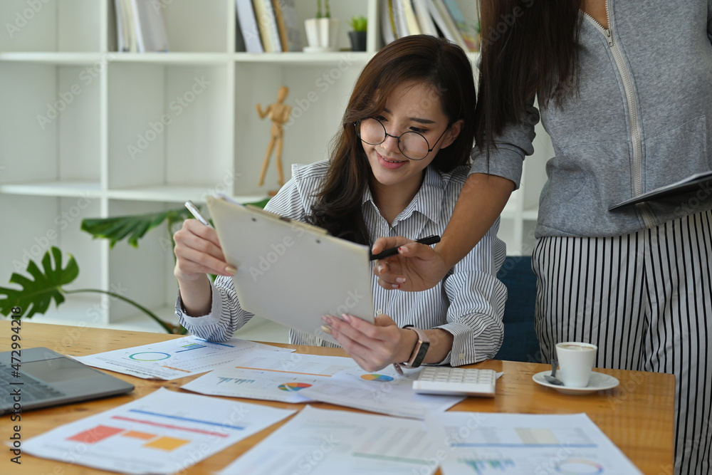 Photo of office women sitting and standing while working together at the wooden working desk surrounded by a computer laptop, document and coffee cup.
