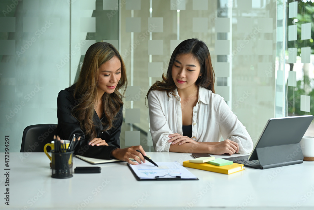 Photo of two female designers working together at the white working desk surrounded by clipboard, digital tablet and office equipment. Photo of two female designers working together at the white working desk surrounded by clipboard, digital tablet and office equipment.