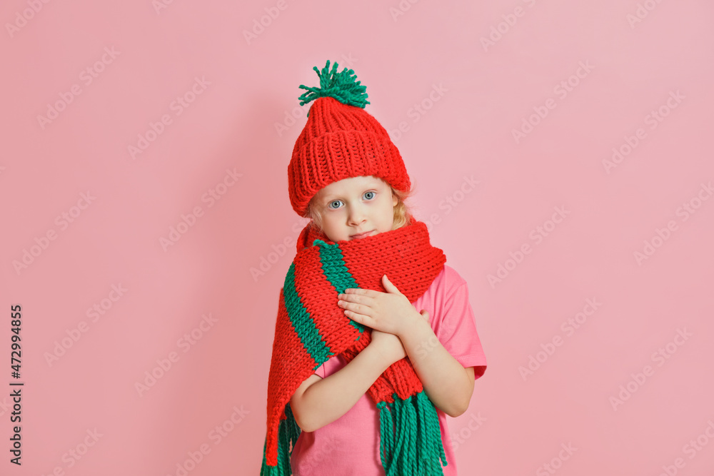 caucasian toddler girl in red knitted scarf and hat on pink background, christmas mood concept copy space