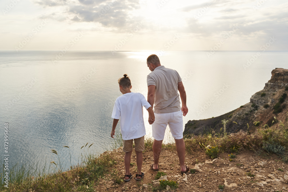 Fototapeta premium the boy holds his dad's hand and they look at the bottom of the cliff against the backdrop of a beautiful rocky sea coast going into the distance