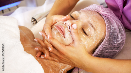 Cosmetologist applies a moisturizing mask on female face. Woman in a spa salon on cosmetic procedures for facial care. White woman getting beauty treatment therapy.