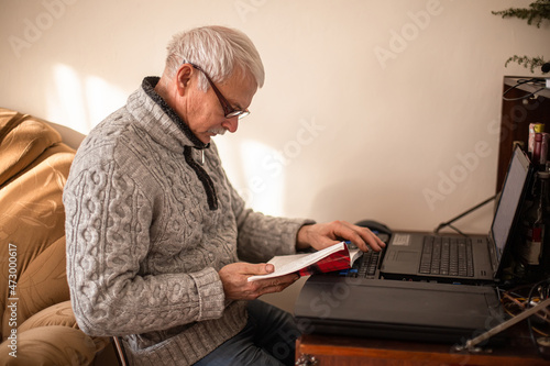 white-haired elderly man reads a book sitting near a table with a laptop. old man reads the instructions for a modern gadget at home