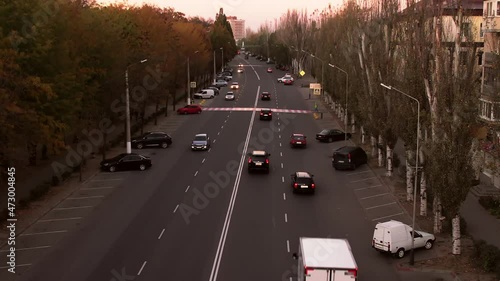Cars drive fast on long wide street marked road with two-lane traffic between colorful trees in city center at sunset aerial view photo