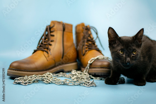 A black cat sits to the right of a pair of new boots against a blue background. Fashionable concept.