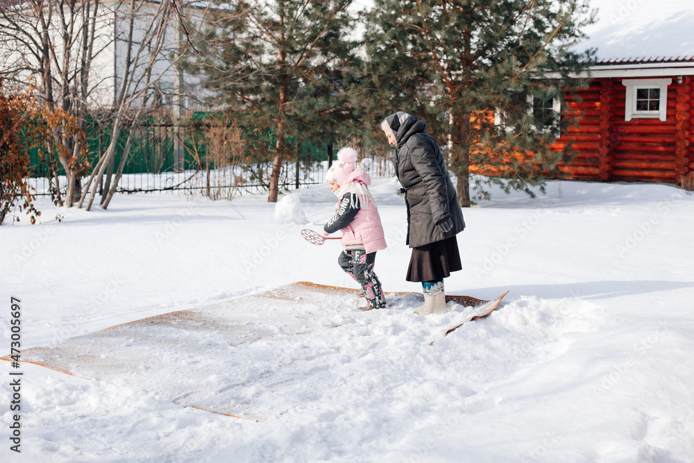 Family is cleaning carpet in backyard. Caucasian woman and child traditionally clean carpet with fresh snow and carpet beater near wooden cottage in winter, side view.