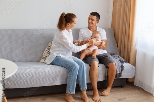 Portrait of delighted young couple wearing casual style clothing sitting on sofa in light living room with infant baby, enjoying to spend together, happy weekend. © sementsova321