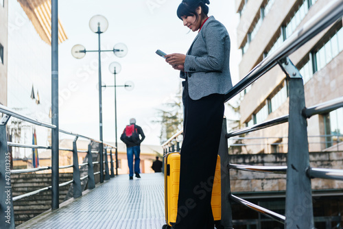 latin business woman with yellow suitcase and using her mobile phone leaning on a railing in the street. business trip concept.