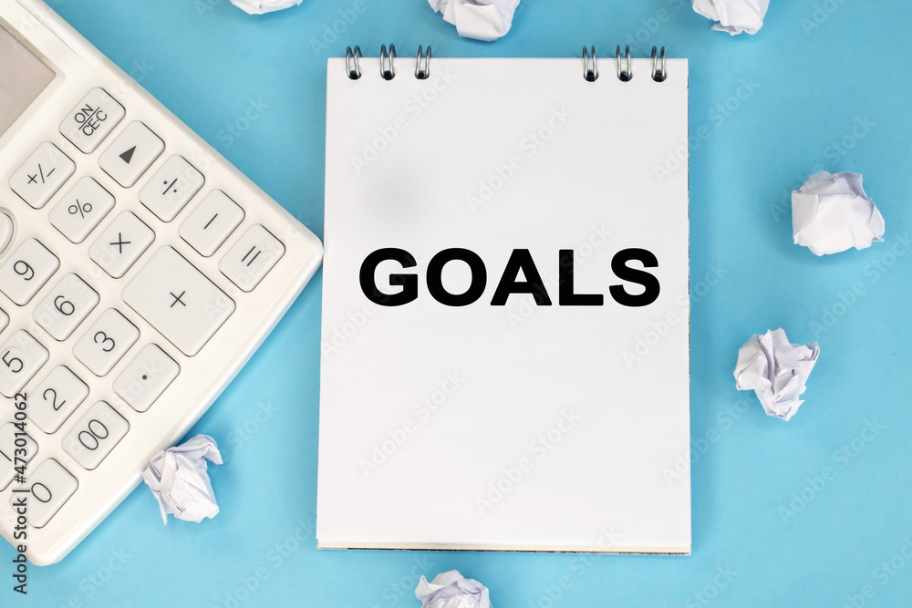 The word goal is written on a notepad. White calculator and crumpled lichty papers on a blue background. The word goal is written on a notepad. White calculator and crumpled lichty papers on a blue background.