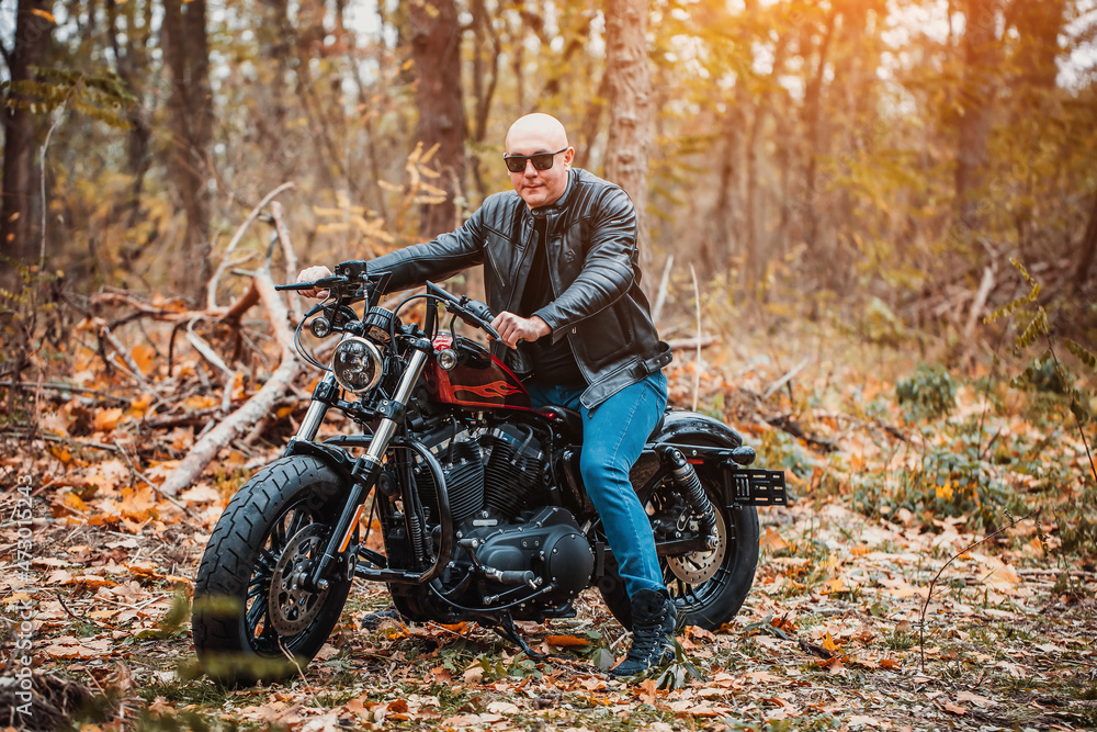 Bald brutal man in sunglasses and a leather jacket sits on a motorcycle on the road in the autumn forest