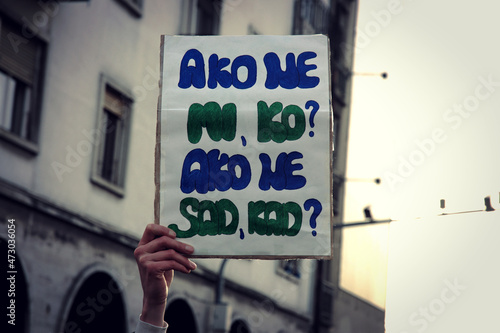 A woman holds a cardboard poster in hand at a protest on the street in Serbia. On it written, if not we, who, if not now, when? photo