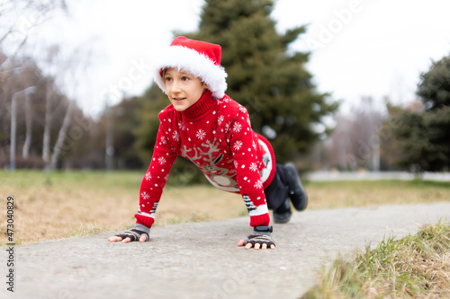 a boy in a warm Christmas sweater with a New Year's deer and a Santa hat got ready to perform a push-up exercise in a city park on the tracks