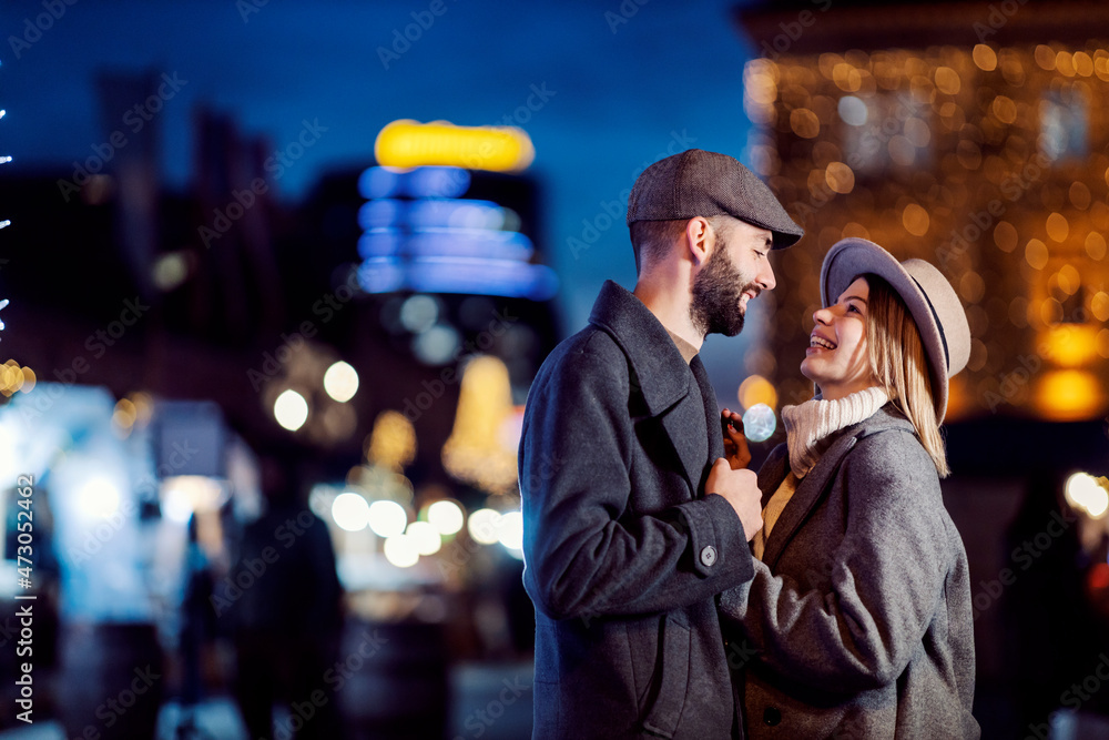 Obraz premium Romantic couple holding hands on the street on Valentine's day. A young happy couple standing on the street, holding hands and making eye contact at night on Valentine's day.