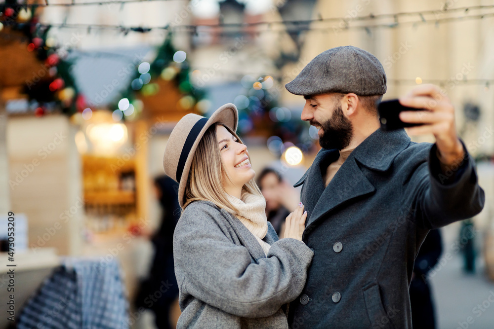 Christmas couple taking selfies outdoors on New Year. A young happy couple standing on the street, laughing, hugging, and taking selfies. Christmas couple taking selfies outdoors on New Year. A young happy couple standing on the street, laughing, hugging, and taking selfies.
