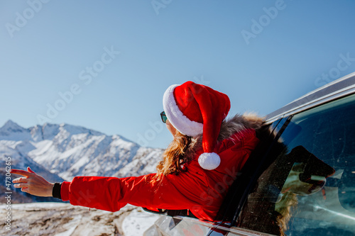 Woman in red winter overalls and Santa cap looking out the car's window and enjoying snowing mountains view. Focus is at the hat. Copy space.