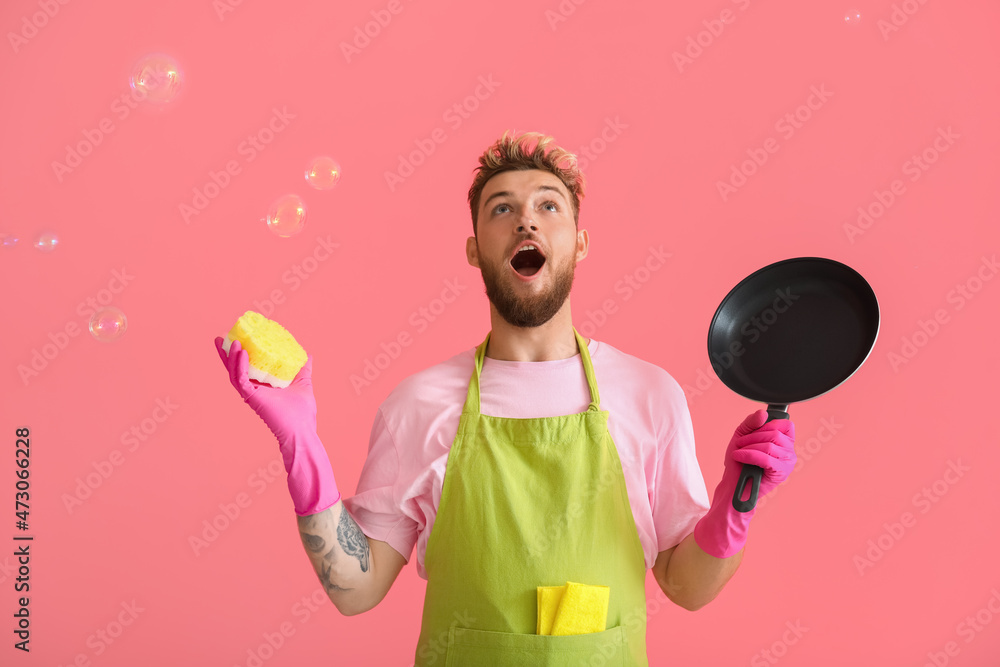 Surprised young man with cleaning sponge and frying pan on pink background Surprised young man with cleaning sponge and frying pan on pink background