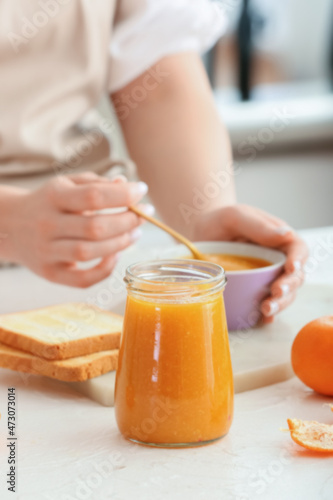 Woman with spoon of tasty tangerine jam at table in kitchen, closeup