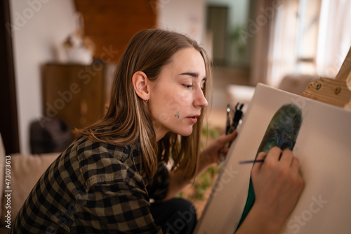One young beautiful caucasian woman artist sitting on the bed sofa at home painting on the canvas holding paintbrush art and creativity professional or hobby concept real people side view copy space