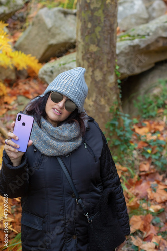 portrait of a woman with sunglasses and wool hat taking self-portraits with smartphone in the forest on an autumn day 
