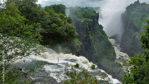 The Zambezi River flows into the gorge  bubbling and foaming. The rapid flow of water between the steep slopes. Fog over the abyss. Lush green vegetation around. Victoria Falls. Zimbabwe.