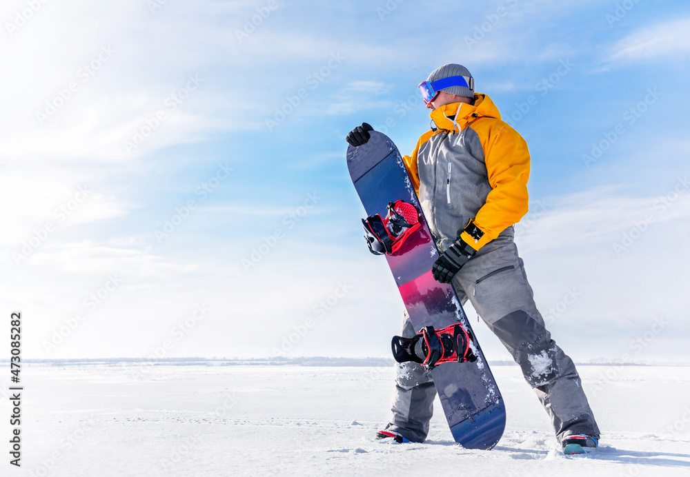 Fototapeta premium Young man holds a snowboard in his hands against the background of a winter landscape. Male in ski goggles and overalls with his board on white snow on a sunny frosty day go in for sports.