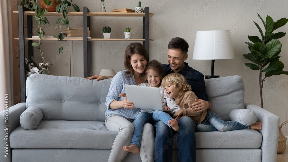 Cheerful millennial parents resting with gen Z kids on couch, making video call, watching movie online, internet TV on laptop computer, having fun, laughing. Happy family enjoying weekend