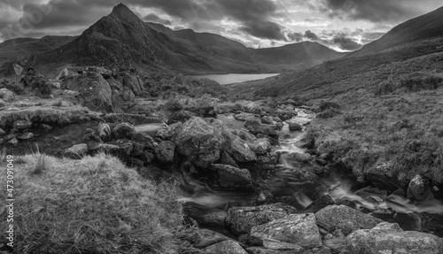 Black and white Epic moody Autumn sunset landscape image of Llyn Ogwen and Tryfan in Snowdonia National Park with stream and rocks in foreground