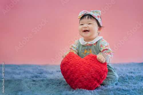 Asian baby valentine with red heart smiling and sitting on pink background. Cute 6 months baby sitting with copy space use as concept of valentine, love, baby or kid department in hospital.