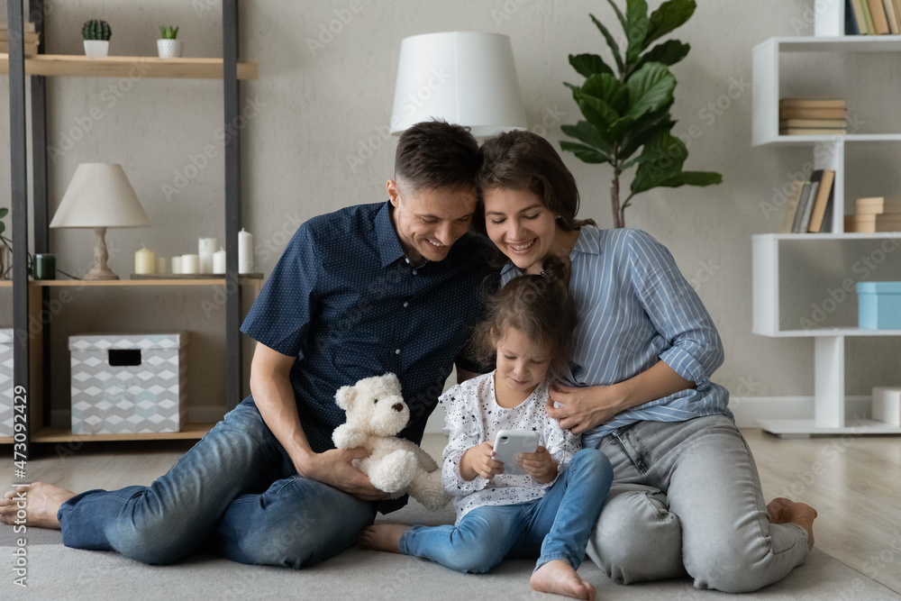 Happy parents hugging cute daughter girl, watching child using smartphone, sitting close on warm carpeted floor, teaching, helping kid to use online app, internet service, read book Happy parents hugging cute daughter girl, watching child using smartphone, sitting close on warm carpeted floor, teaching, helping kid to use online app, internet service, read book