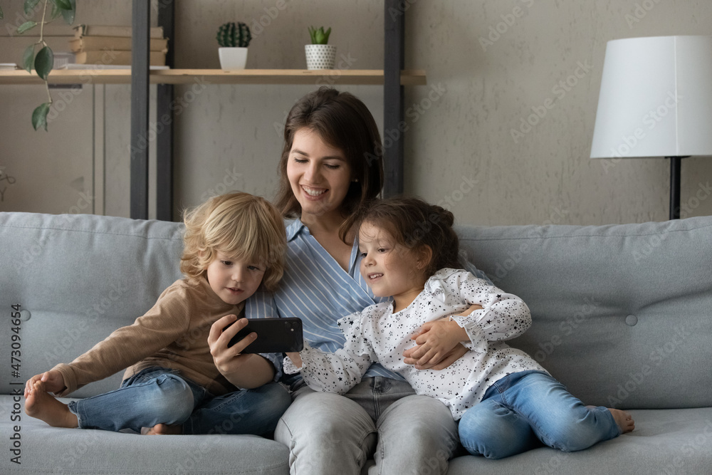 Cheerful mother and two kids taking home selfie on cellphone, talking on video call, resting on sofa in living room together, watching online media, using shopping app on internet