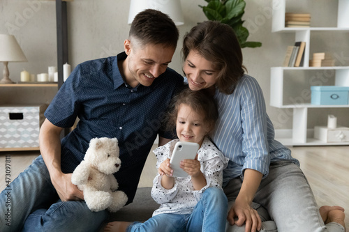Happy laughing parents and adorable preschool kid using smartphone, watching online content, browsing, shopping on internet, talking on video call, taking selfie. Couple keeping parental control