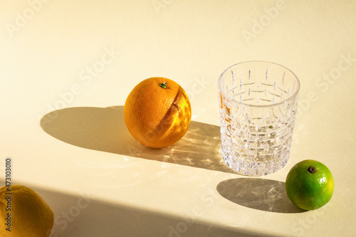 empty crystal glass under hard light, orange's, lime's and lemon's oval shadows, glitter reflection on beige background, food art composition