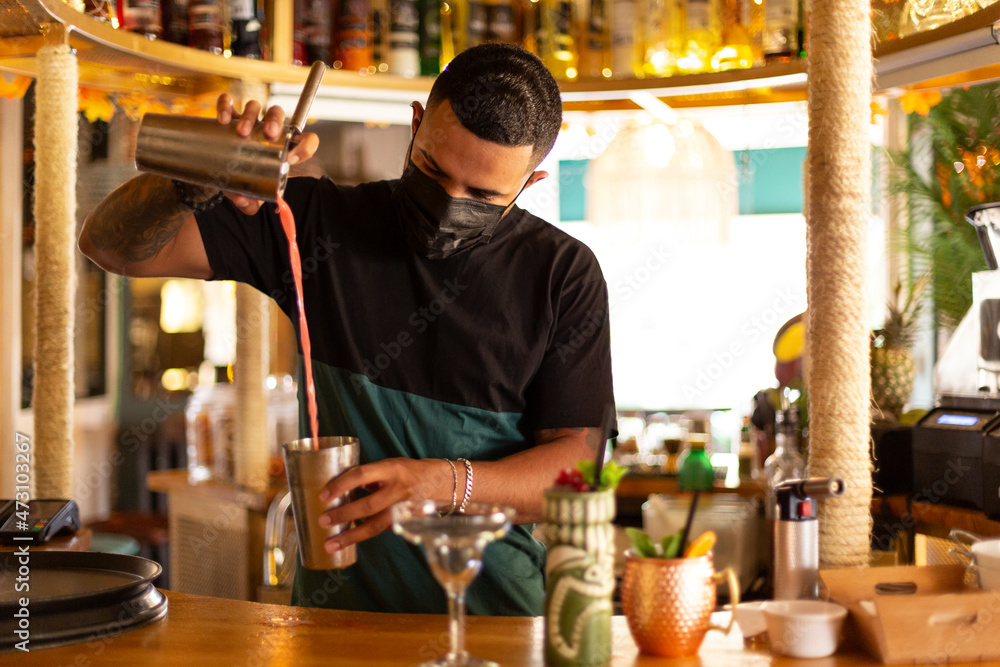 Young Latino bartender preparing a cocktail at the bar. He wears a medical mask.
