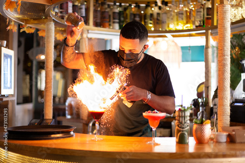 Young latin man preparing an explosive drink at the bar. He is wearing a face mask.