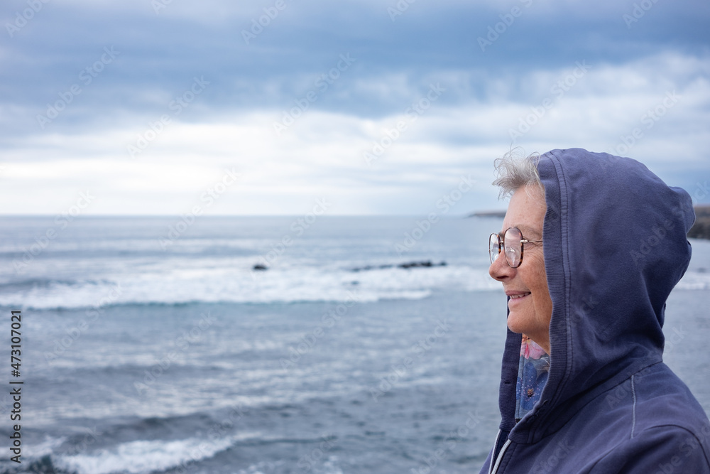 Senior woman with hood on head standing in front to the sea in cloudy day looking away. 70s aged woman enjoying retirement and freedom