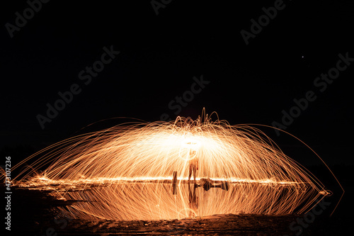 Wide landscape long exposure shot of man spinning glowing steel wool producing light trails. Night photography. Reflection on water surface.