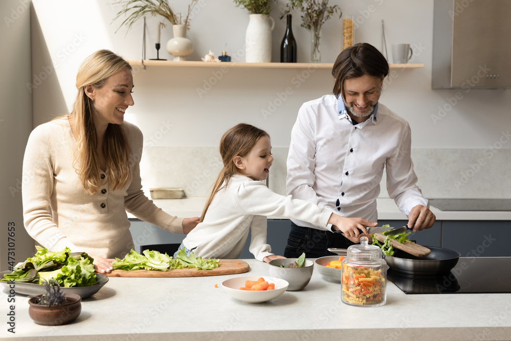 Joyful adorable little preschool kid girl enjoying cooking healthy food with caring parents in modern kitchen. Happy father mother preparing meal with laughing small child daughter on weekend at home.
