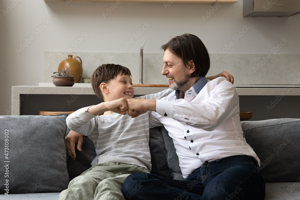 Happy joyful young father and teenage child son bumping fists, enjoying friendly trusted conversation together, sitting on sofa at home. Joyful two male generations family having fun on weekend. Happy joyful young father and teenage child son bumping fists, enjoying friendly trusted conversation together, sitting on sofa at home. Joyful two male generations family having fun on weekend.