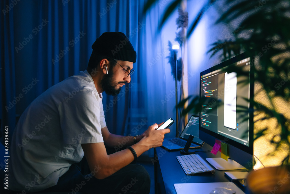 Young man mobile developer writes program code on a computer and check app in smartphone, programmer work in home office. Young man mobile developer writes program code on a computer and check app in smartphone, programmer work in home office.