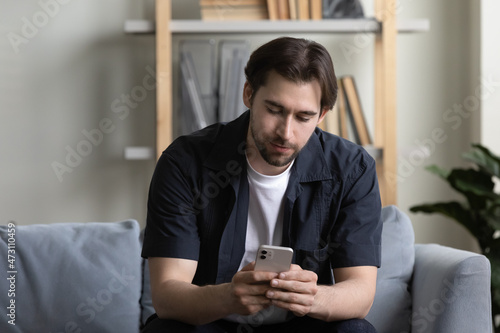 Focused cell user reading text message on mobile phone screen. Young millennial man in casual using online app, banking service on smartphone at home, shopping on internet, chatting virtually