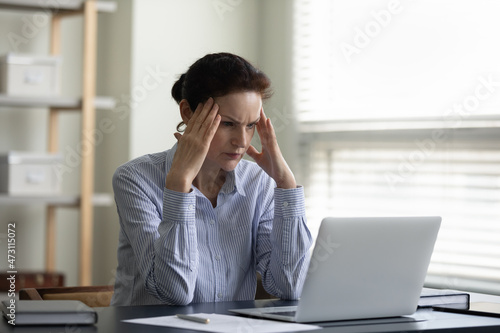 Focused concentrated middle aged businesswoman thinking over problem hard, staring at laptop screen, reading email, touching head, making difficult decision, watching video, webinar, studying online