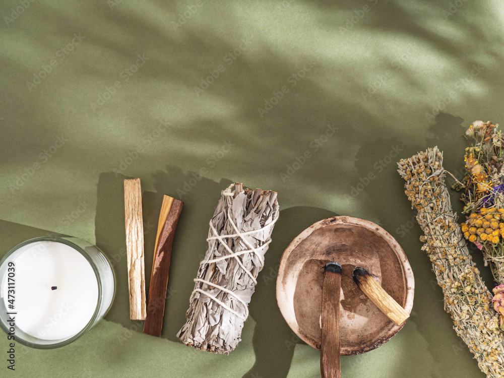 Items for spiritual cleansing - sage and various herbs bundles, palo santo  incense sticks and candle on green background with shadows. Stock Photo