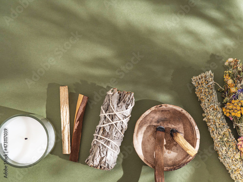 Items for spiritual cleansing - sage and various herbs bundles, palo santo incense sticks and candle on green background with shadows. photo